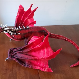 May include: A red dragon plush toy with silver spikes and wings. The dragon has a long tail and is posed with its wings spread out.