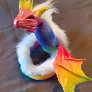 May include: A rainbow-colored dragon figurine with a white fur tail and glittery wings. The dragon has a pink head with a green and yellow crown.