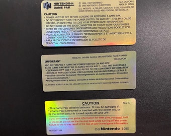 May include: A black and silver Nintendo 64 Game Pak cartridge with a warning label that reads "Caution: This Game Pak contains batteries. It may be damaged if: 1) Game Pak is removed or inserted with the power ON, or 2) the power switch is turned rapidly ON and OFF." The label also includes the text "Nintendo 1985".