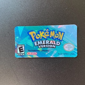 May include: A holographic sticker for the Game Boy Advance video game Pokemon Emerald Version. The sticker features the Pokemon logo, the game title, and the ESRB rating of "Everyone".