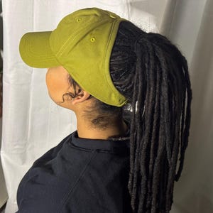 Puede incluir: Una persona lleva una gorra de béisbol verde con una coleta de rastas. La gorra tiene una visera curva y una parte trasera de malla.