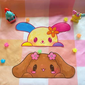 May include: Two cartoon bunny stickers with large ears and smiling faces. One is yellow with a flower, the other is brown with pink flowers. Small star-shaped and toy figures are scattered around the stickers on a pink and orange checkered surface.