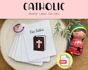 May include: A set of Catholic prayer cards for kids, featuring a white card with the words "Our Father" and an illustration of a closed book with a cross on the cover. The cards are held together with a pink ring and are sitting on a wooden surface. The image also includes a small doll dressed in a red and blue outfit, and a PDF icon.