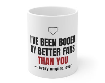 May include: A white ceramic mug featuring a black baseball home plate icon at the top, followed by the black text "I'VE BEEN BOOED BY BETTER FANS", the red text "THAN YOU", and the black text "— every umpire, ever" at the bottom.