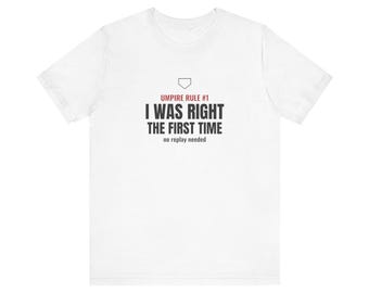 May include: A white short-sleeve t-shirt featuring a black graphic print that reads Umpire Rule #1, I WAS RIGHT THE FIRST TIME, no replay needed, with a small black baseball home plate icon positioned above the text.