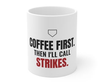 May include: A white ceramic mug featuring a black baseball home plate icon above the black text COFFEE FIRST. THEN I'LL CALL and the red text STRIKES. The mug has a cylindrical shape with a smooth finish and a standard handle on the side.