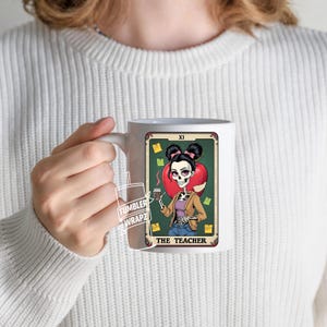 May include: White ceramic mug featuring a tarot card design with a skeleton teacher illustration. The card reads "THE TEACHER" and includes a red apple, coffee, and sticky notes. The mug is held by a person wearing a white sweater.