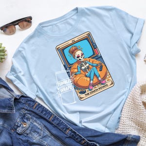 May include: Light blue t-shirt featuring a cartoon skeleton lounging on a beanbag chair in front of a TV. The skeleton is holding a remote control. The text "THE COUCH POTATO" is at the bottom of the image. The shirt also has the text "TUMBLER WRAPZ".