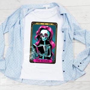 May include: White t-shirt featuring a tarot card design with a skeleton reading a book. The skeleton has pink hair, glasses, and earrings. The card reads "THE READER" in pink text. A light blue button-down shirt is draped over the t-shirt.