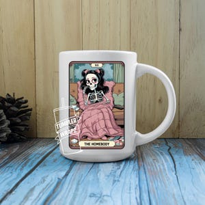 May include: White ceramic mug featuring a tarot card design. The illustration depicts a skeleton figure wrapped in a pink blanket on a couch, with the words "THE HOMEBODY" at the bottom. The mug has a large handle.