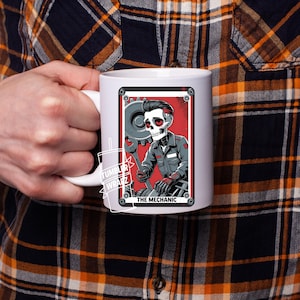 May include: White ceramic mug featuring a stylized illustration of a skeleton mechanic holding a wrench and gear. The design includes a red background and the text "THE MECHANIC". The mug has a white handle.
