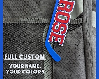 May include: A blue and red plastic hockey stick-shaped luggage tag with the name ROSE printed in bold red letters with a white outline. The text FULL CUSTOM YOUR NAME, YOUR COLORS is visible on the dark gray textured fabric background.