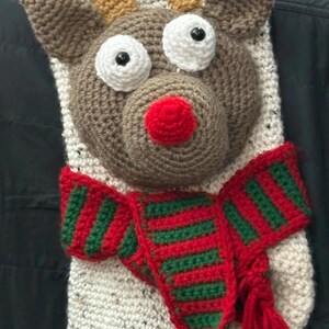May include: A handmade Christmas stocking featuring a crocheted reindeer face. The reindeer has brown antlers, a brown face, white eyes, and a red nose. A red and green striped scarf is around the reindeer's neck.