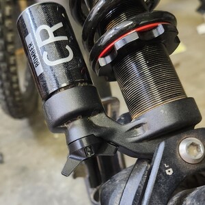 May include: Close-up of a black bicycle suspension fork with a red adjustment knob and a black shock absorber with the text "BOMBER CR" on it.