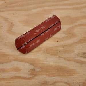May include: A close-up of a black adhesive strip with red 3M VHB tape on both sides. The tape is labeled with the 3M logo and the words "VHB" and "TM".
