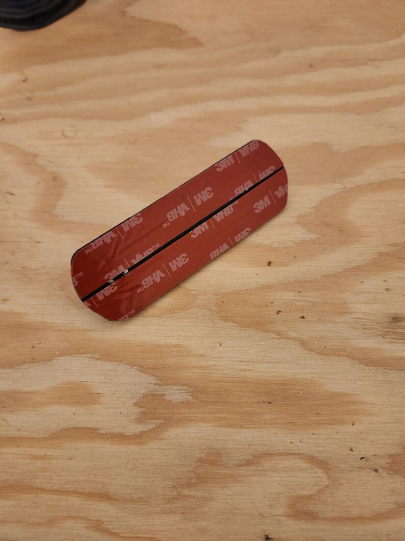 May include: A close-up of a black adhesive strip with red 3M VHB tape on both sides. The tape is labeled with the 3M logo and the words "VHB" and "TM".