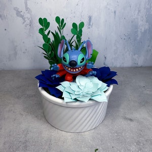 May include: A blue Stitch character figurine sitting in a white planter with green and blue paper succulents.