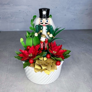 May include: A white ceramic planter with a nutcracker figurine, red poinsettia flowers, and green succulents. The nutcracker is wearing a green and black uniform and holding a drum.
