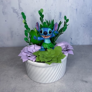 May include: A blue Stitch character figurine is surrounded by purple and green artificial succulents in a white planter. The planter has a textured, swirl design.