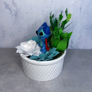 May include: A white ceramic planter with a blue and red Stitch figurine, white paper flowers, and green paper leaves.