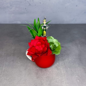 May include: A red ceramic mug with a white rim, filled with a green succulent plant and a red paper flower. A small elf figurine is perched on top of the arrangement.