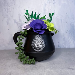 May include: A black cauldron with a white Hogwarts crest, filled with paper flowers in shades of purple, green, and white.