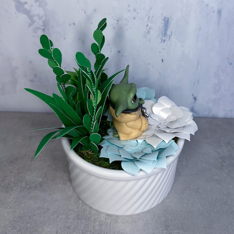 Op de afbeelding: Een klein groen en bruin beeldje van een personage uit de Star Wars-franchise, zittend op een witte pot met blauwe en witte papieren bloemen. De pot is gevuld met groene papieren bladeren.