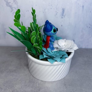 May include: A white ceramic planter with a blue and red Stitch figurine surrounded by green paper leaves and white and blue paper flowers.