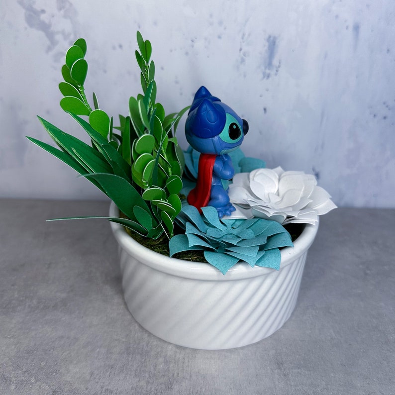 May include: A white ceramic planter with a blue and red Stitch figurine surrounded by green paper leaves and white and blue paper flowers.