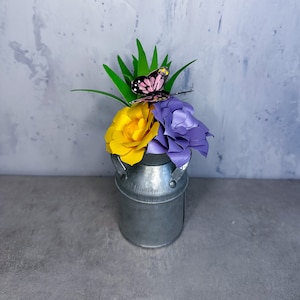 May include: A silver milk can with a yellow and purple paper flower arrangement and a pink butterfly.