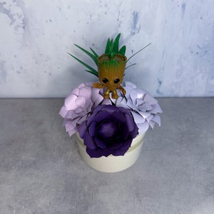 May include: A small brown tree-like character figurine with large black eyes stands atop a cluster of paper flowers. The arrangement features one deep purple flower surrounded by several light lavender paper blooms in a white cylindrical container.