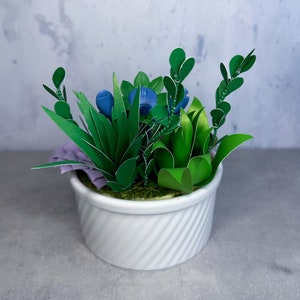 May include: A white ceramic planter with a textured surface holds a variety of green paper plants, including succulents and ferns. The plants are arranged in a natural, organic way, creating a lush and vibrant display.