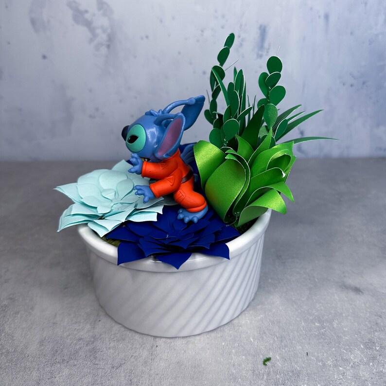 May include: A white ceramic planter with a blue and orange Stitch figurine and green paper succulents.
