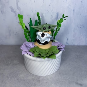 Op de afbeelding: Een klein groen beeldje van een personage uit de Star Wars-franchise, zittend in een klein voertuig, is omringd door kunstbloemen en groen in een witte keramische pot.
