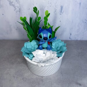 May include: A white ceramic planter with a blue Stitch figurine sitting on top of a bed of green and blue paper succulents.