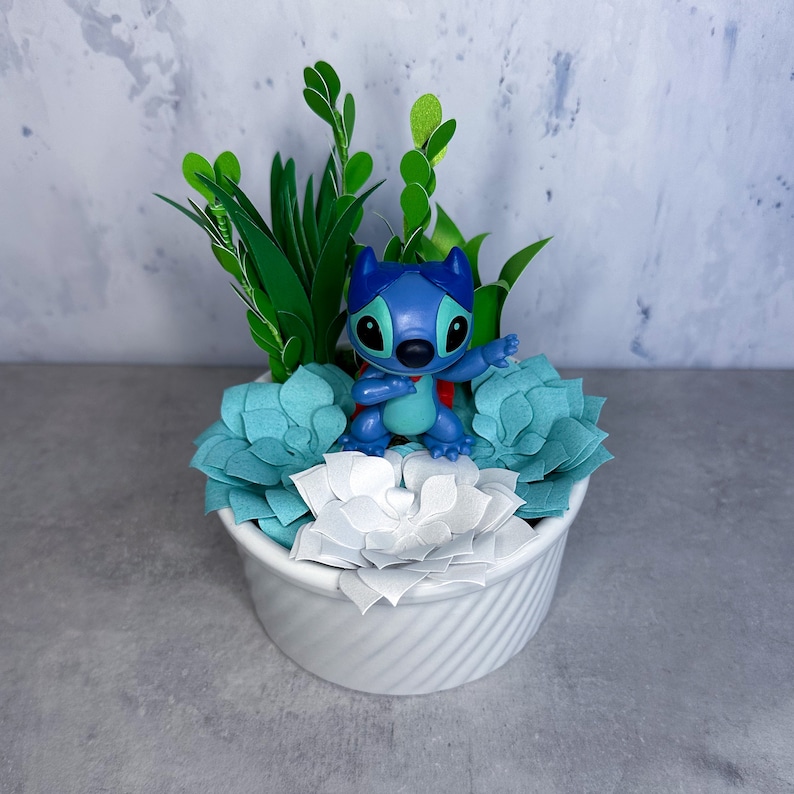 May include: A white ceramic planter with a blue Stitch figurine sitting on top of a bed of green and blue paper succulents.