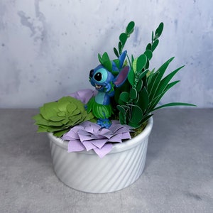 May include: A blue Stitch figurine with green leaves and purple flowers in a white planter. The planter has a swirl design.