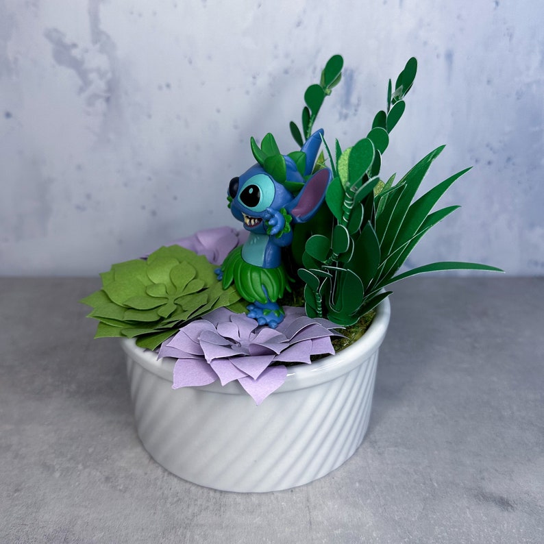 May include: A blue Stitch figurine with green leaves and purple flowers in a white planter. The planter has a swirl design.
