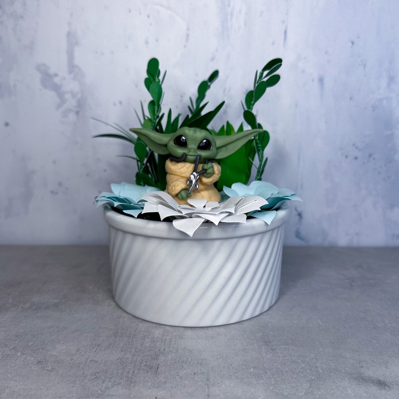 Op de afbeelding: Een klein groen en geel beeldje van een personage uit de Star Wars-franchise, zittend in een witte keramische pot met blauwe en witte papieren bloemen. De pot heeft een swirl-patroon.