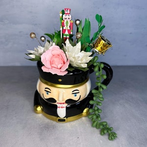 May include: A black ceramic mug with a nutcracker design, decorated with pink and white flowers, green foliage, and a gold-colored ornament.