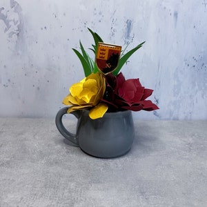May include: A gray ceramic mug containing a bouquet of paper flowers in metallic gold and deep maroon. Green paper leaves are interspersed among the blooms. A small shield-shaped pin with the text Gryffindor is tucked into the floral arrangement.