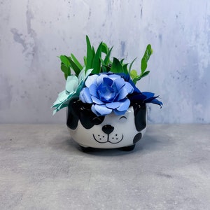 May include: A white ceramic planter shaped like a dog's face with black spots. The planter is filled with a small arrangement of blue and green paper flowers.
