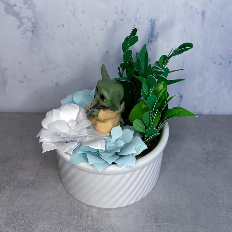 Op de afbeelding: Een witte keramische pot met een groene papieren plant en een klein groen en bruin beeldje. De pot is versierd met witte papieren bloemen.