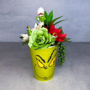 May include: A green Grinch-themed cup with a festive arrangement of succulents, poinsettias, and Christmas-themed decorations.