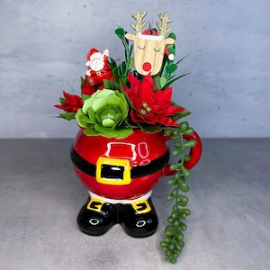 May include: A red ceramic mug shaped like Santa Claus with black boots and a belt, filled with artificial green succulents, red poinsettia flowers, a small Santa figurine, and a wooden reindeer topper with gold glitter antlers.