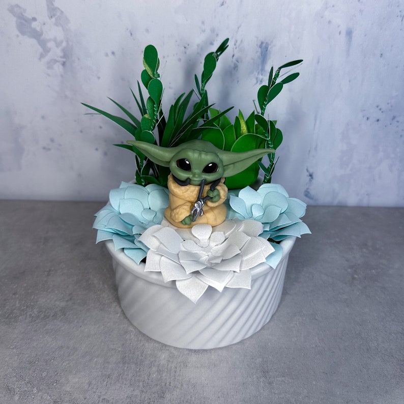 Op de afbeelding: Een klein groen beeldje van een personage uit de Star Wars-franchise, zittend in een witte pot met blauwe en witte papieren bloemen. Het beeldje houdt een klein groen voorwerp in zijn hand.