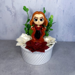 May include: A white ceramic planter with a red and white paper flower arrangement. A small figurine of a red-haired woman in a red and black costume stands in the center of the arrangement.