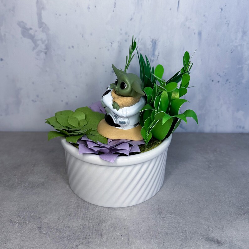Op de afbeelding: Een klein groen beeldje van een personage uit de Star Wars-franchise, zittend in een witte pot met groene en paarse kunstmatige vetplanten.