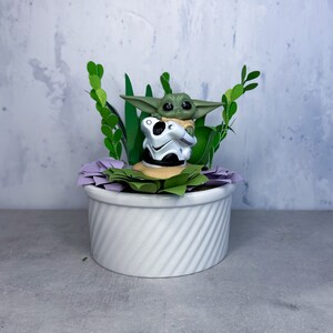 Op de afbeelding: Een klein groen beeldje van een personage uit de Star Wars-franchise, zittend op een bed van paarse en groene papieren bloemen in een witte keramische pot. Het beeldje houdt een klein wit voorwerp vast.