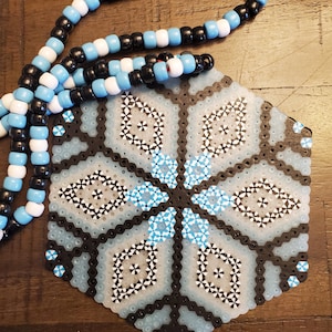Può includere: Un mandala esagonale di perline con un motivo a fiocco di neve. Il mandala è realizzato con perline nere, bianche e blu chiaro. Una collana di perline nera e bianca è appoggiata su una superficie di legno.
